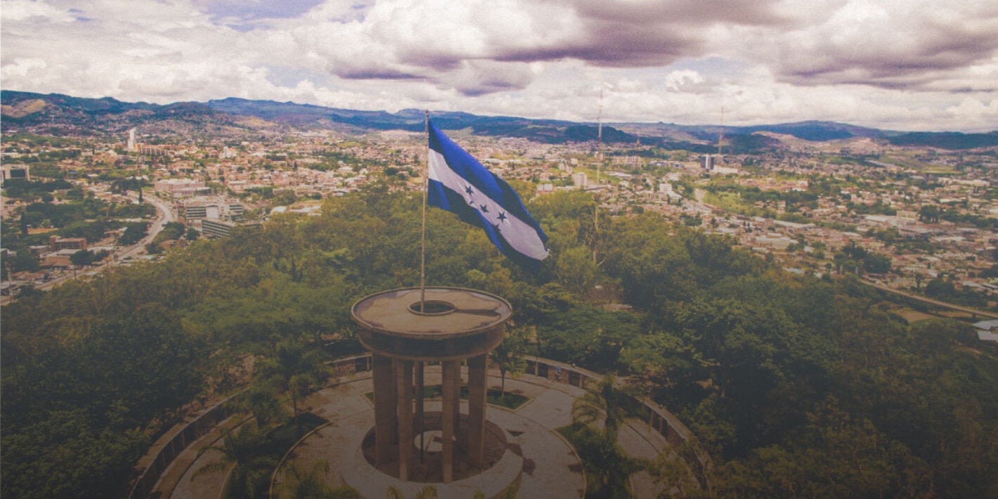 Foto de un paisaje con la bandera de Honduras en primer plano ondeando desde lo alto de una colina, montada sobre una columna circular. Al fondo, panorámica de la ciudad de Tegucigalpa y en el horizonte una línea azul de montañas