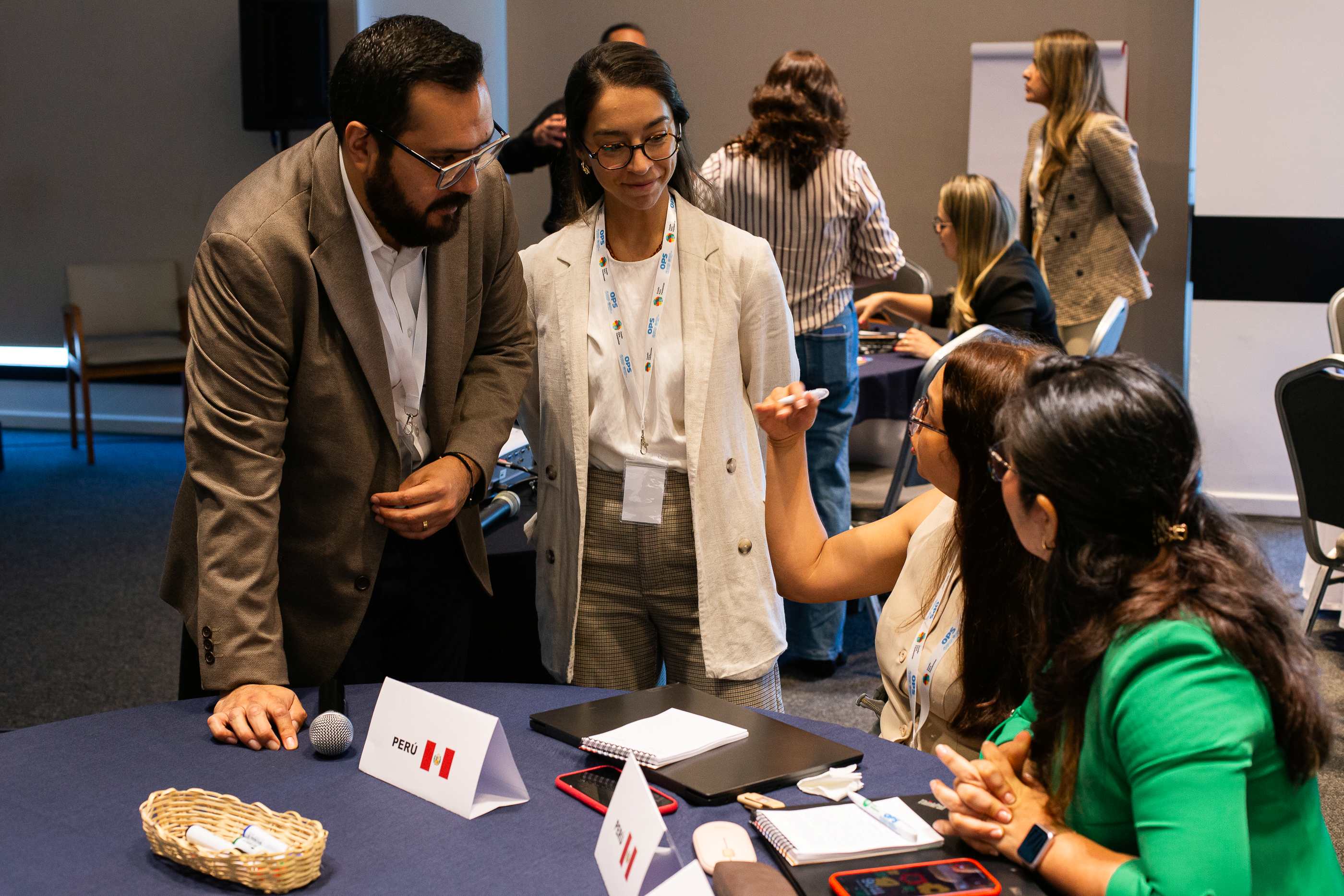Participantes de evento conversando en un salon de eventos. Dos personas paradas conversan con dos personas sentadas en mesa de evento.