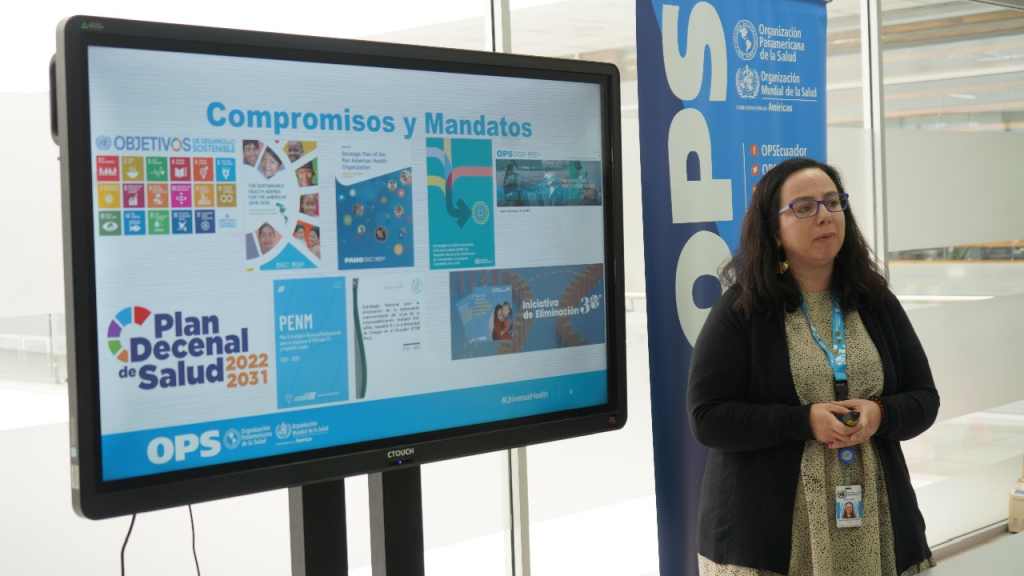 Una mujer, quien expone, está al lado de un monitor que dice Compromisos y Mandatos