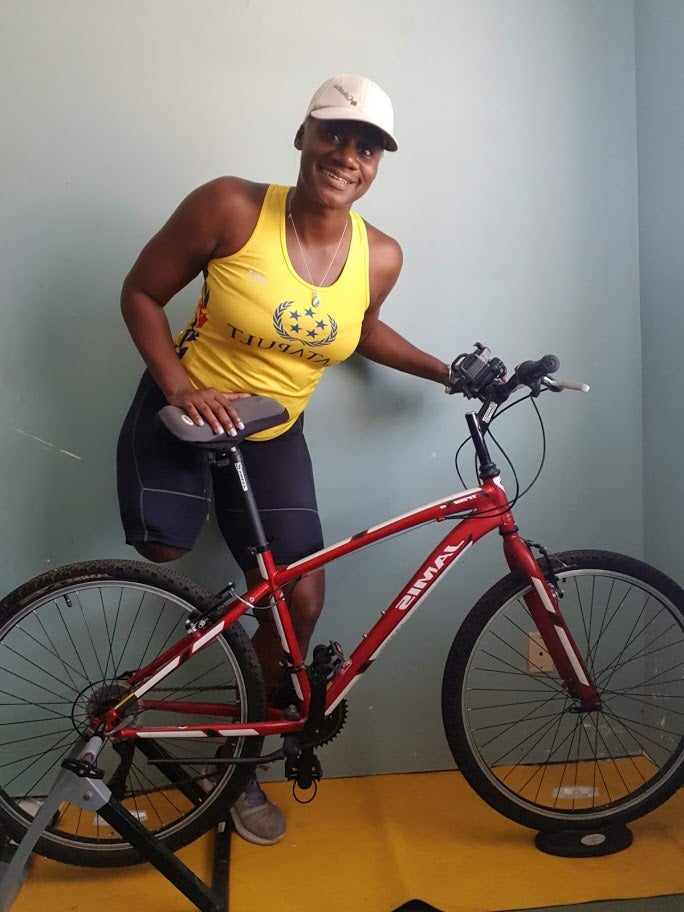A limb difference, black female wearing a tan hat, yellow Catapult shirt and black cyclist tights, holding on to her cycling bike with is red frame with white writing one a yellow workout mat.