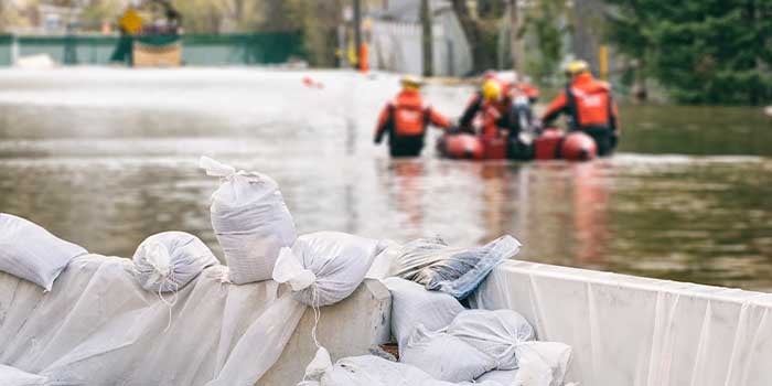 A flood barrier with sandbags is visible in the foreground. Behind it is extensive flooding with flooded homes. A rescue team wearing orange waterproof clothing and yellow helmets can be seen standing in the middle of the flood pushing a red inflatable rescue raft