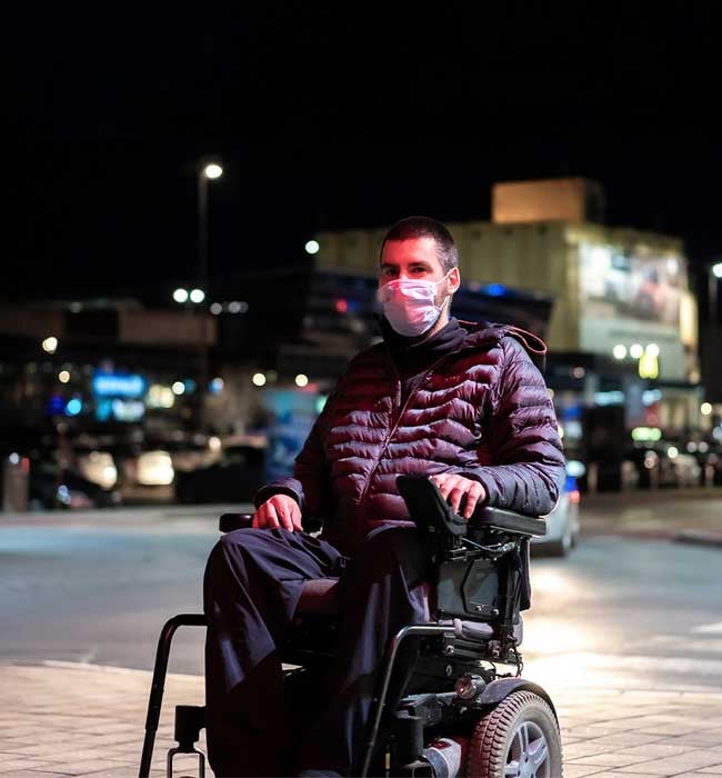 A man wearing a black coat and black pants and a white mask is in a power wheelchair crossing a crosswalk. His left hand is on the armrest/control knob. It is night time with buildings and city lights in the background.