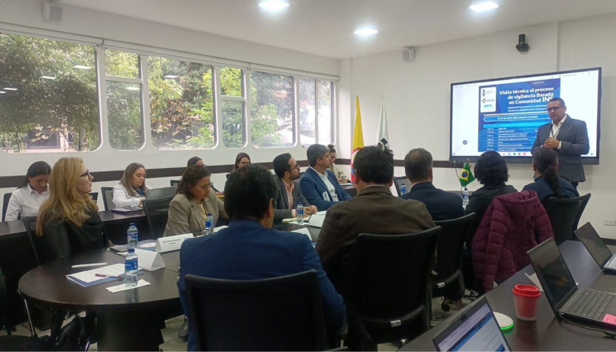 ejecutivos sentados en mesa de salón de reuniones. Uno de los participantes está parado al frente, junto a una pizarra, exponiendo
