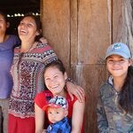 Cuatro mujeres sonríen en la puerta de una cabaña, tienen edades diferentes. La tercera mujer sostiene un niño de unos 2 años en su regazo