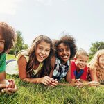Fotografía de seis niños y niñas de edad preadolescente de diferente origen étnico, que formando una fila están tumbados con el vientre sobre la hierba y el torso levantado y los rostros sonrientes