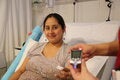 Pregnant woman sitting on a hospital bed holding a piece of cotton after a blood sample, while another hand in the foreground displays a blood glucose meter
