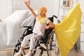 A child sitting in a wheelchair in a bright bedroom raises one arm in a triumphant pose, wearing a yellow cape and awareness ribbon, symbolizing strength, hope, and resilience during childhood cancer treatment.