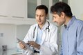 Male doctor with a white coat shows a clipboard to a male patient dressed with a blue shirt.