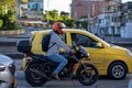 Motociclista con casco rojo, circulando en una avenida urbana, con un vehículo amarillo a su lado izquierdo