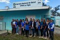 Grupo de trabajadores de salud del centro de atención primaria La Vigía y expertos de OPS posan frente al centro de salud, que tiene las paredes pintadas de verde aguamarina, con un cielo azul con nubes dispersas y un frente de hierba