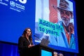 Woman on podium giving speech with presentation on a big screen behind her, with the Decade of Healthy Aging in the Americas card with slogan that reads "Adding life to years"