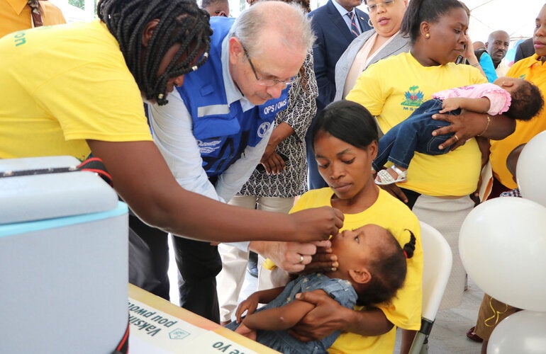 Le Représentant de l'OPS/OMS en Haïti, le Dr Luis Felipe Codina administre un vaccin Polio Oral à un enfant sous le regard de la Ministre de la Sante Publique