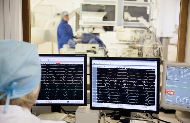 Photo of the back of the head of a healthworker, behing him three screens showing paralel lines that indicates the heart function. Behind the screens there is a big window leading to a room with health equipment where another health worker is accompanying a patient laid on the machine