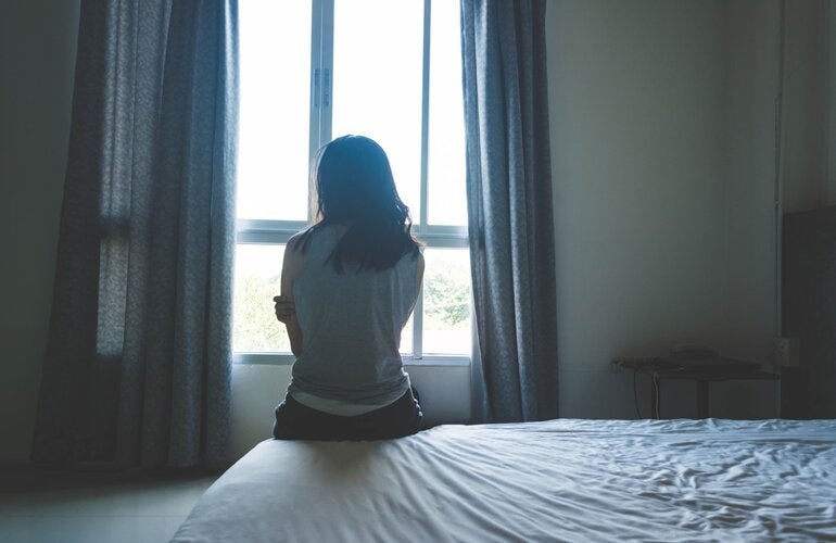 Mujer en la penumbra de una habitación, sentada sobre una cama de espaldas a la cámara, mirando hacia una ventana