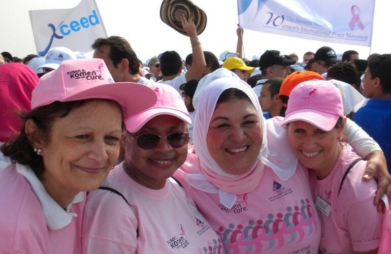 A group of four women dressed in pink t shirts embrace each other smiling. In the background other people is gathering with some banners on a breast cancer awarenes event