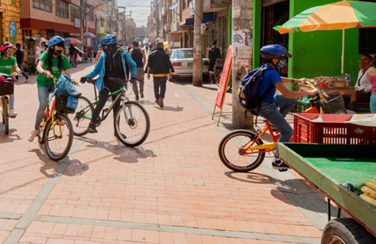 Foto de um grupo de jovens com capacetes de bicicleta e andando de bicicleta em uma rua urbana ensolarada com uma banca de frutas na frente