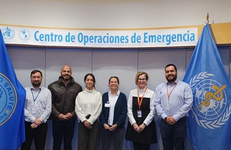 Personas (hombres y mujeres) posan a la cámara en el Centro de Operaciones de Emergencias de la OPS. A ambos lados, están las banderas de la OPS y la OMS.