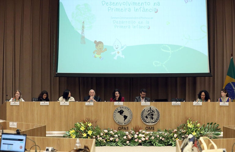 Mesa de abertura do evento com oito pessoas e um telão ao fundo escrito "Desenvolvimento na primeira infância"