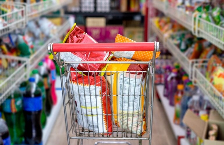 Photo of a supermarket cart filled with huge bags of chips, in the middle of two supermarket aisles with more ultraprocessed products