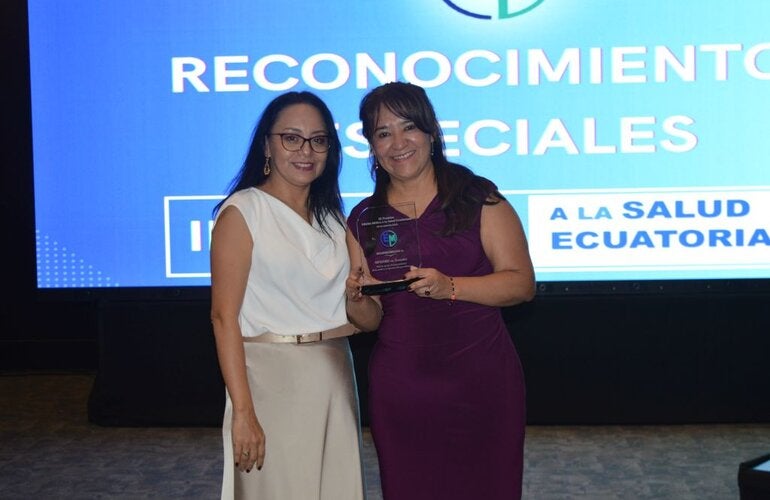 La representante de la OPS/OMS en Ecuador, Dra. Sonia Quezada, recibe de la periodista Cristina Coello, el reconocimiento entregado a la oficina de parte de Edición Médica.