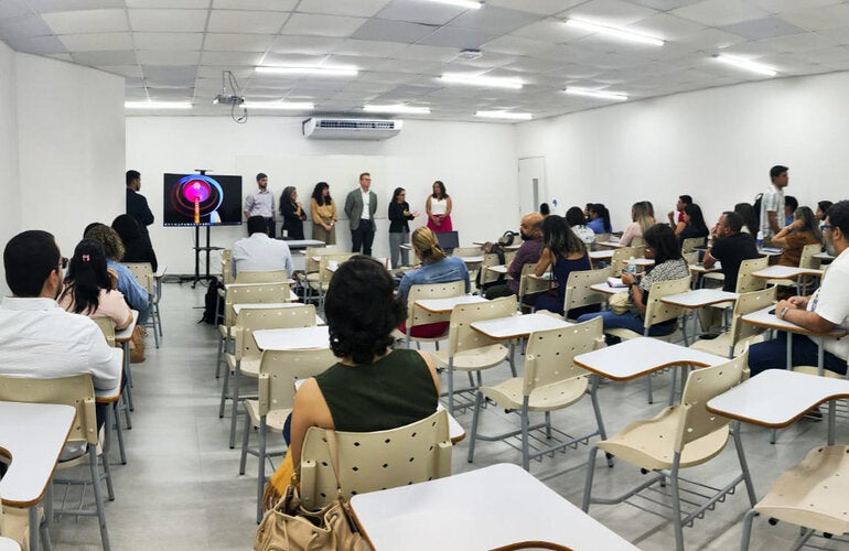 Profissionais sentados em mesas de sala de aula, ouvindo palestra da equipe da OPAS e parceiros