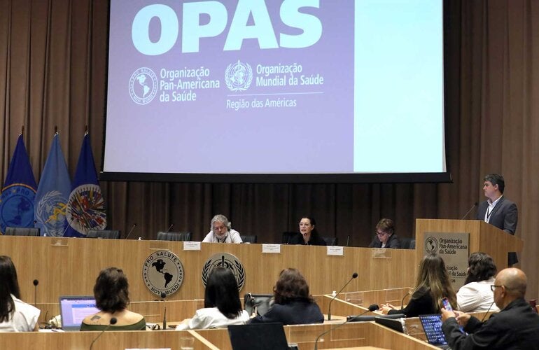 Mesa de abertura do evento, com telão ao fundo com a logo da OPAS