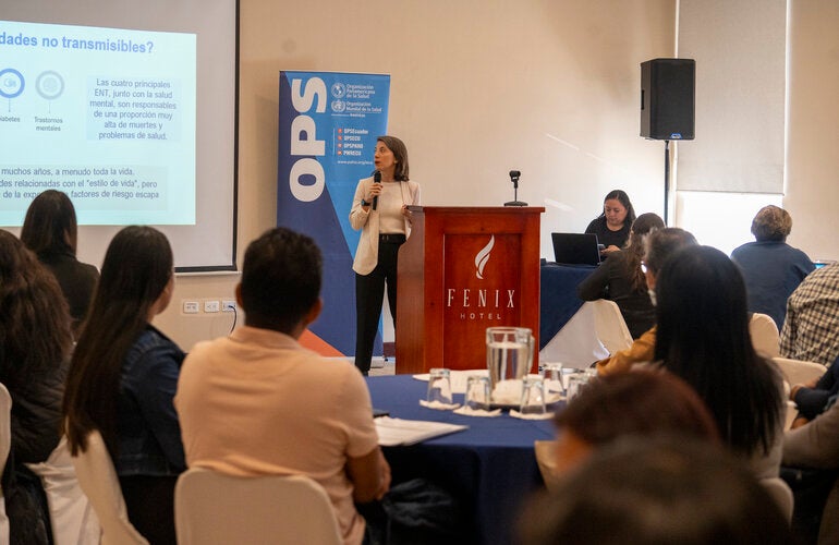 Sesión de entrenamiento de la encuesta STEPS en Quito, Ecuador (8–10 de septiembre de 2025), con una persona presentando frente a un atril del Fenix Hotel, acompañada de un banner de la OPS. Se proyecta una diapositiva sobre enfermedades no transmisibles, mientras la audiencia escucha desde mesas redondas con manteles azules.