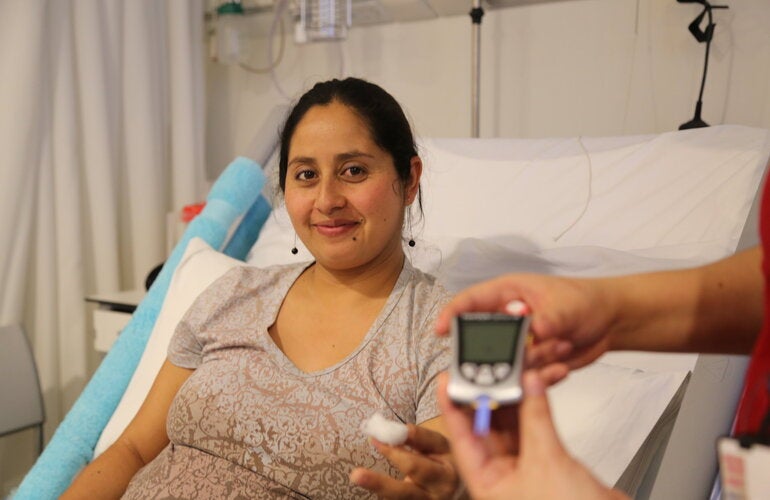 Pregnant woman sitting on a hospital bed holding a piece of cotton after a blood sample, while another hand in the foreground displays a blood glucose meter