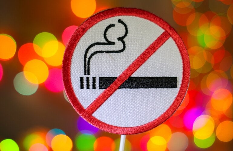 Panneau rond à bordure rouge avec une cigarette noire et de la fumée barrée par une ligne diagonale rouge, indiquant interdiction de fumer. Arrière-plan flou avec des lumières colorées.