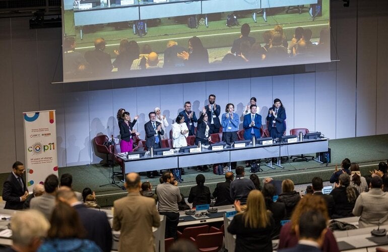 Conference room at COP11 meeting; panelists stand and applaud at the front, audience members also standing and clapping.
