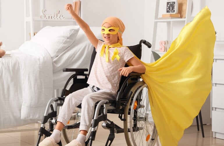 A child sitting in a wheelchair in a bright bedroom raises one arm in a triumphant pose, wearing a yellow cape and awareness ribbon, symbolizing strength, hope, and resilience during childhood cancer treatment.