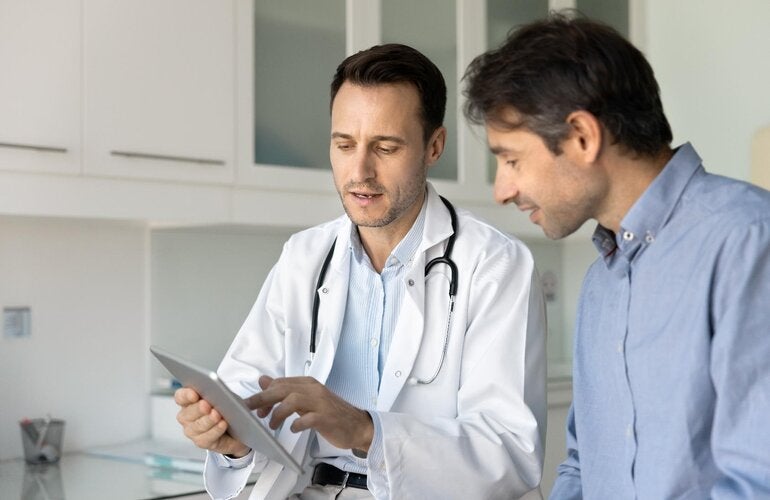 Male doctor with a white coat shows a clipboard to a male patient dressed with a blue shirt.