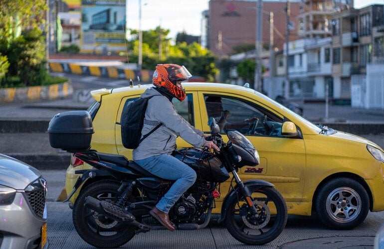 Motociclista con casco rojo, circulando en una avenida urbana, con un vehículo amarillo a su lado izquierdo