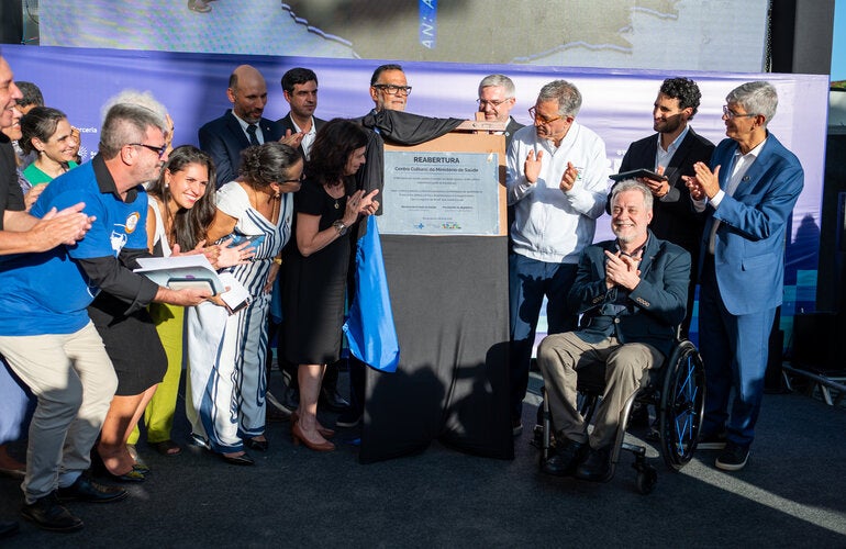 Grupo de autoridades, pesquisadores e participantes reunidos ao redor da placa de inauguração durante a cerimônia de reabertura do Centro Cultural do Ministério da Saúde, no Rio de Janeiro. Algumas pessoas aplaudem enquanto a placa comemorativa é descerrada; em primeiro plano, um homem em cadeira de rodas sorri e participa do momento.