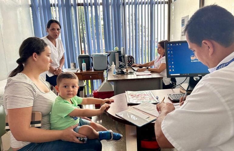 Adult holding a small child during a consultation at a health center, while healthcare staff assist and enter information on a computer.