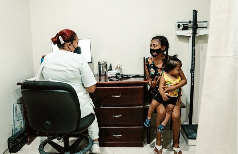 Health personnel seated in a consultation room speaks with an adult holding a young child in their arms.