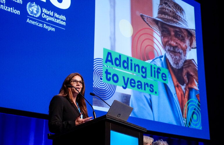 Woman on podium giving speech with presentation on a big screen behind her, with the Decade of Healthy Aging in the Americas card with slogan that reads "Adding life to years"