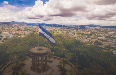 Foto de un paisaje con la bandera de Honduras en primer plano ondeando desde lo alto de una colina, montada sobre una columna circular. Al fondo, panorámica de la ciudad de Tegucigalpa y en el horizonte una línea azul de montañas