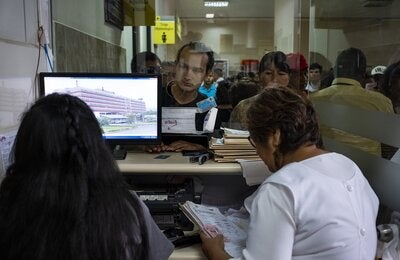 Image of the registration area in a hospital, where two health workers are attending a line of people in front of her
