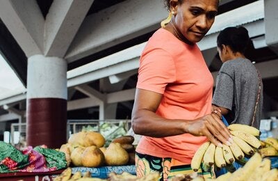 Photo of a middle aged woman dressed with an orange t-shirt grabbing a bunch of bananas on a market fruit stall
