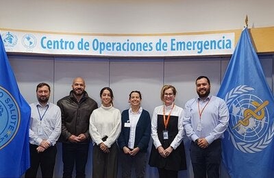Personas (hombres y mujeres) posan a la cámara en el Centro de Operaciones de Emergencias de la OPS. A ambos lados, están las banderas de la OPS y la OMS.