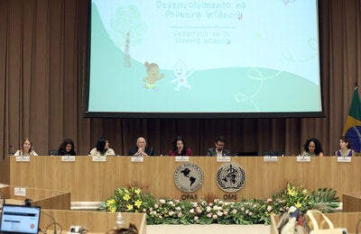 Mesa de abertura do evento com oito pessoas e um telão ao fundo escrito "Desenvolvimento na primeira infância"