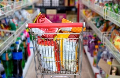 Photo of a supermarket cart filled with huge bags of chips, in the middle of two supermarket aisles with more ultraprocessed products
