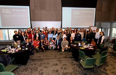 Group photo during the Regional Consultation for the Prioritization of TCIM Research in the Americas, held June 11-12, 2025, in São Paulo, Brazil. Approximately 50 participants pose smiling in the center of a room with tables and working materials. In the background, two screens show the agenda for the “TM Themes and Priorities” session.