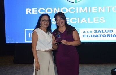 La representante de la OPS/OMS en Ecuador, Dra. Sonia Quezada, recibe de la periodista Cristina Coello, el reconocimiento entregado a la oficina de parte de Edición Médica.