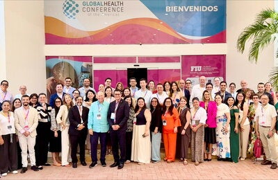 Los participantes del taller “Evidencia y herramientas disponibles para la eliminación del cáncer cervicouterino en las Américas” posan para una foto grupal frente a la entrada de un edificio. En el fondo, se puede ver un cartel que dice "GLOBAL HEALTH CONFERENCE of the Americas" y "BIENVENIDOS". También hay logotipos de FIU (Florida International University) y otros elementos decorativos. Los asistentes están vestidos con ropa formal y llevan gafetes colgados al cuello.