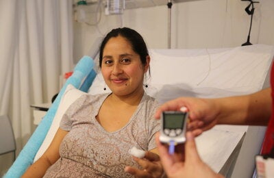 Pregnant woman sitting on a hospital bed holding a piece of cotton after a blood sample, while another hand in the foreground displays a blood glucose meter