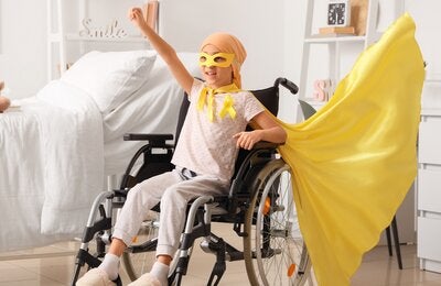 A child sitting in a wheelchair in a bright bedroom raises one arm in a triumphant pose, wearing a yellow cape and awareness ribbon, symbolizing strength, hope, and resilience during childhood cancer treatment.