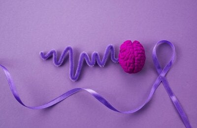 Sobre fondo morado, una ilustración de un cerebro en rosa fucsia del que sale una línea morada hacia la izquierda, al lado derecho un lazo de tela morado