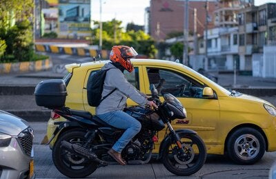 Motociclista con casco rojo, circulando en una avenida urbana, con un vehículo amarillo a su lado izquierdo